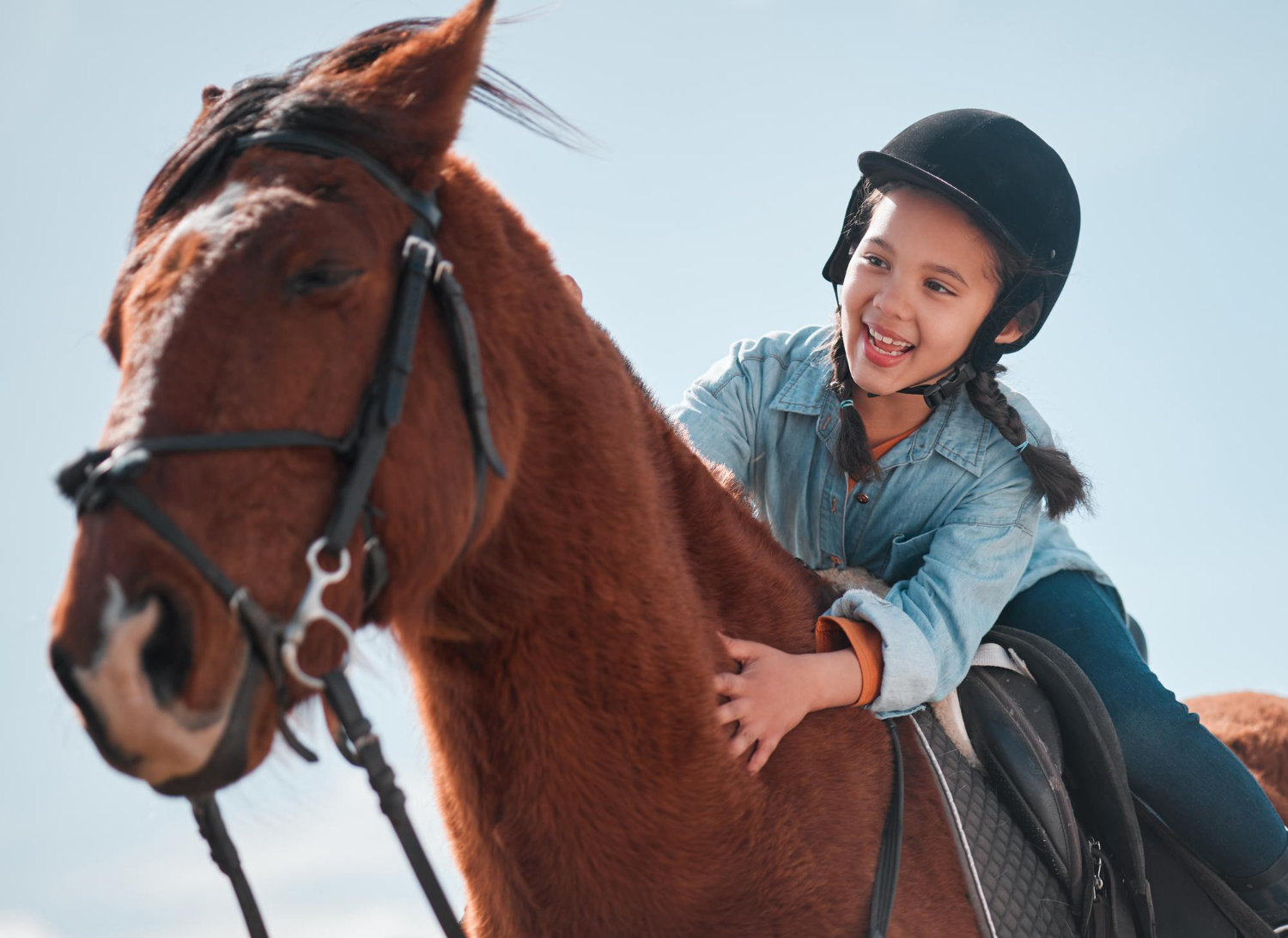 Girl with braided hair smiles while riding a brown horse against a clear blue sky.